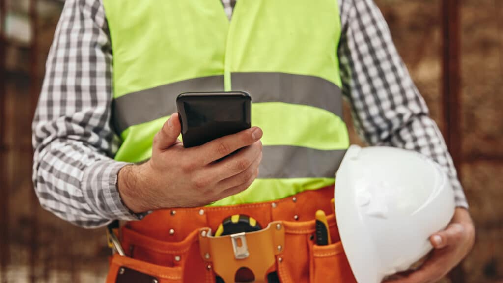 Construction project manager in safety vest using a smartphone on-site, holding a hard hat representing digital tools and hands-on oversight in multifamily development.